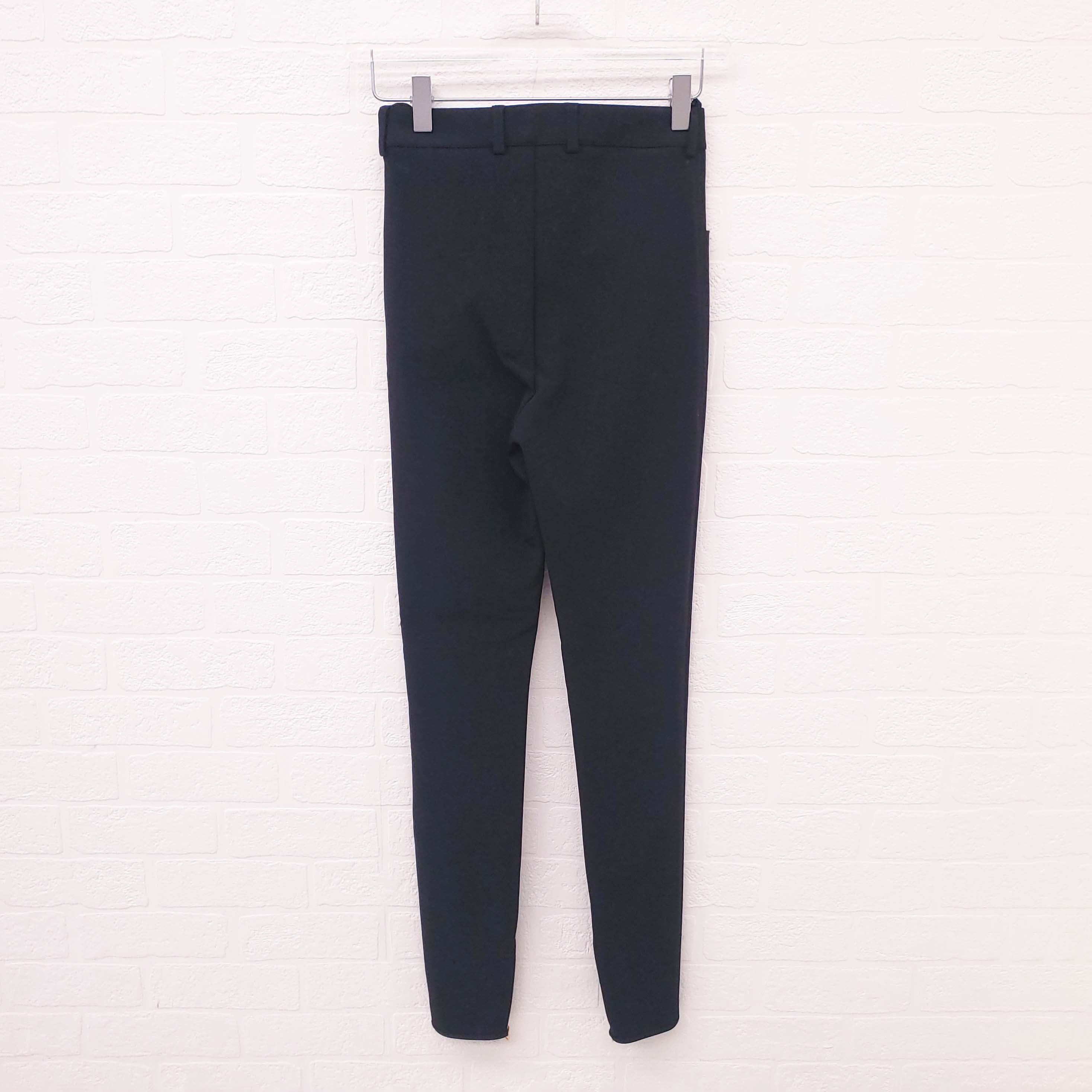 Black Celine Runway pants on a white hanger against a white brick wall background.